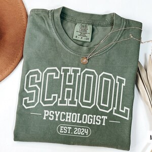 May include: A green t-shirt with white text that reads "SCHOOL PSYCHOLOGIST EST. 2024".