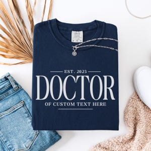 May include: Navy blue t-shirt with white text that reads "EST. 2025 DOCTOR OF CUSTOM TEXT HERE".