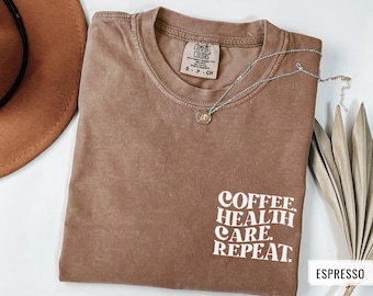 May include: A brown t-shirt with the text "COFFEE. HEALTH CARE. REPEAT." printed in white. The shirt is made of a soft, comfortable material and has a relaxed fit.
