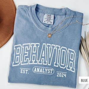 May include: A blue jean t-shirt with the text "BEHAVIOR ANALYST 2024" printed on the front. The shirt is made of a soft, comfortable fabric and has a relaxed fit.