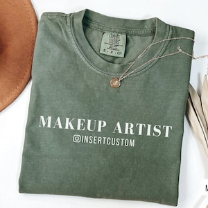 May include: A green t-shirt with the text "MAKEUP ARTIST" and "@INSERTCUSTOM" in white lettering. The shirt is folded and laid flat on a white surface.