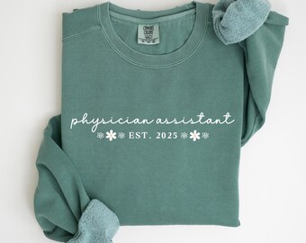 May include: A green sweatshirt with the text "physician assistant est. 2025" in white with small white flowers on either side of the text.
