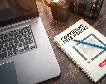Può includere: Un quaderno bianco con una rilegatura a spirale su un tavolo di legno. Il quaderno ha il testo "COPYRIGHT FREE IMAGES 2018 GUIDE" sulla copertina. Una matita blu è appoggiata sul quaderno. Un laptop e una fotocamera sono anche sul tavolo.
