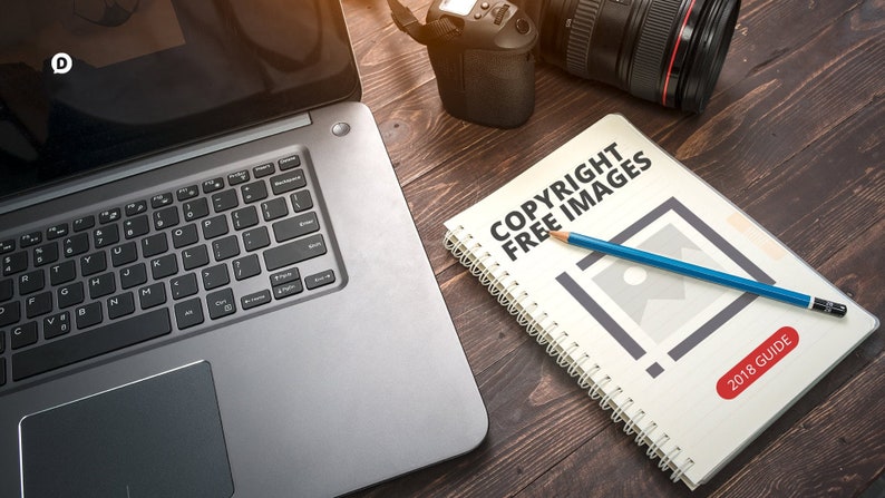 May include: A white notebook with a spiral binding on a wooden table. The notebook has the text "COPYRIGHT FREE IMAGES 2018 GUIDE" on the cover. A blue pencil is lying on top of the notebook. A laptop and a camera are also on the table.
