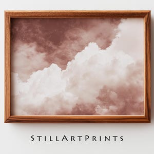 May include: A framed print featuring a cloudscape in shades of mauve and white. The clouds are fluffy and textured, set against a darker mauve background. The wooden frame adds a warm, natural touch. The text "STILLARTPRINTS" is at the bottom.