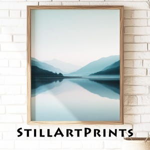 May include: A framed print of a tranquil lake scene with mountains in shades of blue and teal. The water reflects the landscape, creating a mirrored effect. The text "STILLARTPRINTS" is visible at the bottom of the print, which is in a wooden frame.
