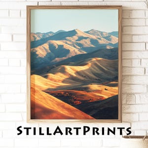 Puede incluir: Una impresión enmarcada de un paisaje montañoso, que representa colinas y picos dorados y azules, iluminados por el sol. La imagen tiene un cielo despejado y el texto "STILLARTPRINTS" en la parte inferior. La impresión está en un marco de madera.