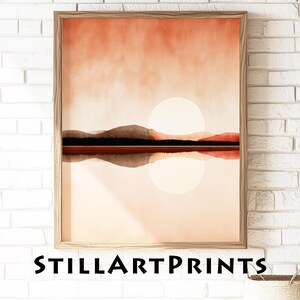 Puede incluir: Una impresión artística enmarcada de un paisaje con una puesta de sol sobre un lago. La obra de arte tiene un estilo minimalista con tonos cálidos de naranja, marrón y beige. El texto "STILLARTPRINTS" está impreso en la parte inferior del marco.