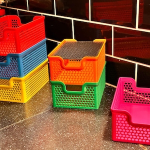 Könnte beinhalten: Eine Sammlung farbenfroher Kunststoff-Aufbewahrungskörbe in Rot, Blau, Gelb, Orange, Grün und Pink. Die Körbe haben ein perforiertes Design und sind gestapelt oder einzeln platziert. Ideal zum Organisieren von Gegenständen.