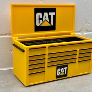 May include: Yellow metal toolbox with the CAT logo on the lid and front. The toolbox has a hinged lid and multiple drawers.