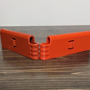 Pu&ograve; includere: Due oggetti rettangolari di plastica arancione brillante con un ritaglio a forma di I e una serie di creste verticali. Gli oggetti sono posizionati ad angolo, creando una forma a V. Gli articoli sono su una superficie di legno.