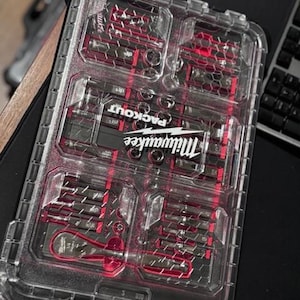 Könnte beinhalten: Transparenter Milwaukee Packout Werkzeugorganizer mit roten Akzenten. Das Gehäuse ist in mehrere Fächer unterteilt, die jeweils verschiedene Werkzeuge enthalten. Das Milwaukee-Logo und "PACKOUT" sind sichtbar.