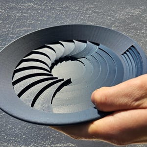 Puede incluir: Un objeto circular gris oscuro con un diseño en espiral. El objeto se sostiene en una mano y parece estar hecho de un material sólido. El diseño en espiral tiene una serie de líneas curvas y paralelas.