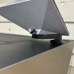 Könnte beinhalten: Ein graues Solarpanel mit einer schwarzen Halterung. Das Panel ist nach oben geneigt und an einer grauen Oberfläche befestigt.