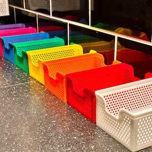 May include: A row of colorful plastic storage bins in various colors, including purple, pink, blue, green, yellow, orange, red, and white. The bins have a perforated design and are arranged in a line.
