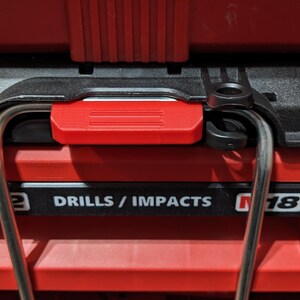 Pu&ograve; includere: Primo piano di una cassetta degli attrezzi rossa con manico nero e meccanismo di bloccaggio rosso. La cassetta riporta la scritta "M12 DRILLS / IMPACTS M18". Un manico argentato si incurva sopra.