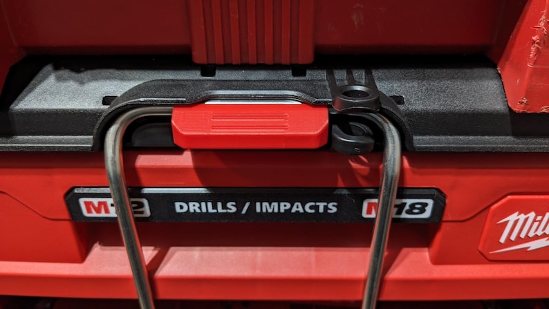 Pu&ograve; includere: Primo piano di una cassetta degli attrezzi rossa con manico nero e meccanismo di bloccaggio rosso. La cassetta riporta la scritta "M12 DRILLS / IMPACTS M18". Un manico argentato si incurva sopra.