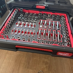 Könnte beinhalten: Eine schwarze und rote Milwaukee Packout Werkzeugkasten-Schublade, gefüllt mit verschiedenen Steckschlüsseln, Verlängerungen und Ratschen. Die Werkzeuge sind ordentlich in einer roten Einlage angeordnet, mit dem Milwaukee-Logo auf der Vorderseite der Schublade.