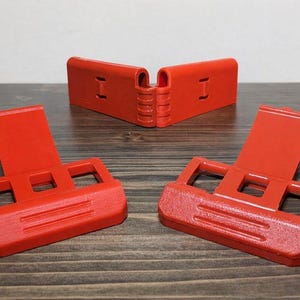 Pu&ograve; includere: Tre oggetti di plastica rosso brillante sono esposti su una superficie di legno. Due sono angolati verso l'osservatore, mentre un terzo &egrave; posizionato sullo sfondo. Ogni oggetto ha una base rettangolare con ritagli quadrati e un pannello verticale incernierato.