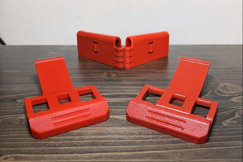 Pu&ograve; includere: Tre oggetti di plastica rosso brillante sono esposti su una superficie di legno. Due sono angolati verso l'osservatore, mentre un terzo &egrave; posizionato sullo sfondo. Ogni oggetto ha una base rettangolare con ritagli quadrati e un pannello verticale incernierato.