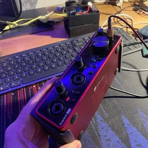 Pode incluir: Interface de áudio Scarlett 2i2 vermelha com botões pretos e conectores de entrada. O dispositivo possui uma tomada para fone de ouvido e vários botões de controle. A interface é conectada a um computador por meio de um cabo. Projetado para gravação e produção de áudio.