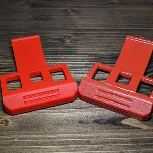 Pu&ograve; includere: Due supporti per telefono in plastica rosso brillante sono esposti su una superficie di legno. Ogni supporto ha una base rettangolare con tre ritagli quadrati e un supporto verticale. I supporti sono progettati per tenere un telefono in posizione verticale.