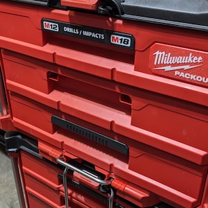Pu&ograve; includere: Cassette degli attrezzi Packout Milwaukee rosse e nere impilate. La cassetta degli attrezzi superiore ha un coperchio nero con una maniglia rossa. Il testo "M12 DRILLS / IMPACTS M18" &egrave; visibile sul davanti.