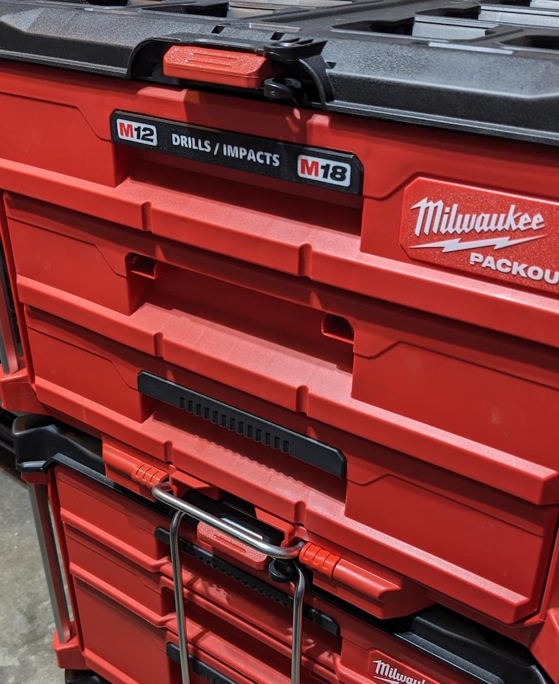 Pu&ograve; includere: Cassette degli attrezzi Packout Milwaukee rosse e nere impilate. La cassetta degli attrezzi superiore ha un coperchio nero con una maniglia rossa. Il testo "M12 DRILLS / IMPACTS M18" &egrave; visibile sul davanti.