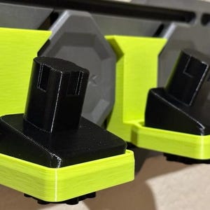 Może przedstawiać: Dwa czarne uchwyty na baterie z neonowo-zieloną podstawą, przeznaczone do akumulatorów elektronarzędzi. Uchwyty są zamontowane na szarej ścianie. Obraz jest dobrze oświetlony i pokazuje szczegóły projektu drukowanego w 3D.