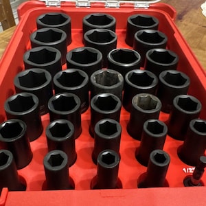 Könnte beinhalten: Eine rote Werkzeugkiste gefüllt mit einem Satz schwarzer Schlagschraubernüsse. Die Nüsse sind in Reihen und Spalten angeordnet, in verschiedenen Größen. Die Werkzeugkiste ist geöffnet und zeigt die geordnete Anordnung der Werkzeuge.