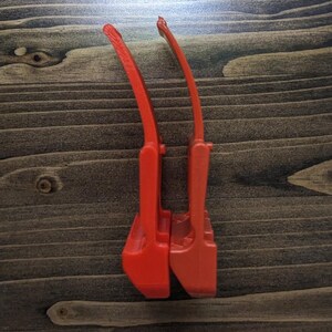 Pu&ograve; includere: Due utensili in plastica rosso vivo, forse per un compito specifico, sono posizionati verticalmente su uno sfondo di venature del legno scuro. Gli utensili hanno un manico curvo e una base rettangolare.