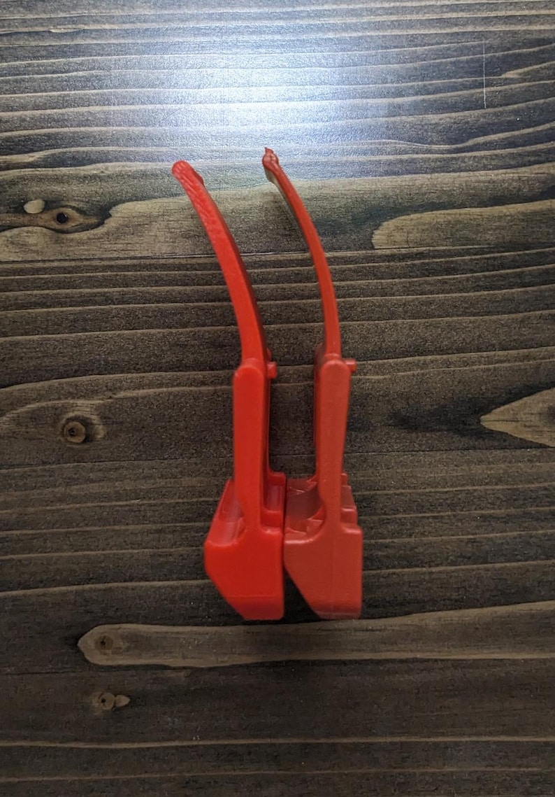 Pu&ograve; includere: Due utensili in plastica rosso vivo, forse per un compito specifico, sono posizionati verticalmente su uno sfondo di venature del legno scuro. Gli utensili hanno un manico curvo e una base rettangolare.