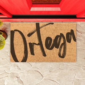 May include: A brown coir doormat with the name "Ortega" written in black brush script.