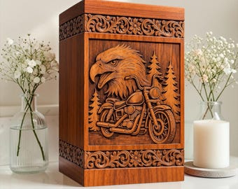 Könnte beinhalten: Eine rechteckige Urne aus Holz mit einer detaillierten Reliefschnitzerei eines Adlerkopfes über einem Motorrad vor einem Waldhintergrund. Die oberen und unteren Ränder sind mit komplizierten floralen Rankenmustern verziert.