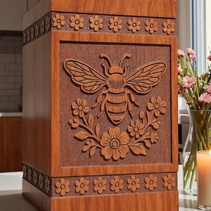 May include: A wooden box with a carved bee and floral design. The box is a warm brown color, with a rectangular shape. The bee is centered on the front panel, surrounded by flowers and leaves. Floral patterns also adorn the top and bottom edges.