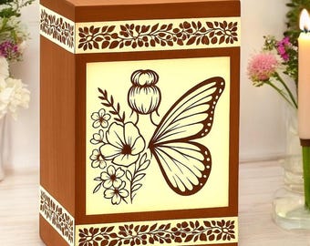Puede incluir: Una urna funeraria rectangular de madera con un panel central claro que presenta un gráfico marrón oscuro de una persona con un moño, un ala de mariposa grande y flores. Los bordes superior e inferior tienen cenefas de hojas.
