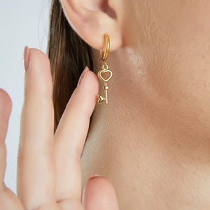 May include: Gold hoop earrings with a heart-shaped key charm dangling from the bottom.