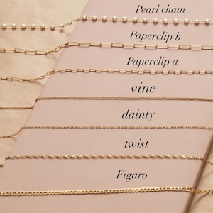 May include: A collection of gold necklaces displayed on a light pink surface. The necklaces include a pearl chain, paperclip chains, a vine chain, a dainty chain, a twist chain, and a Figaro chain. Each chain is labeled with its name in a handwritten font.