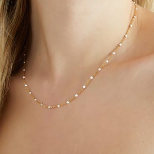 May include: Delicate gold necklace featuring small, evenly spaced white beads. The necklace is displayed against a skin tone background, highlighting its fine details and elegant design. A simple and classic piece of jewelry.