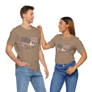 May include: Brown t-shirts with a graphic of an American flag and a duck wearing sunglasses. The text "Ameri-Quack" is below the duck and flag. The t-shirts are worn by a man and a woman.