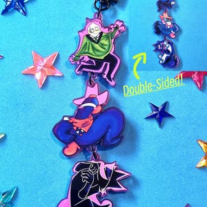 Puede incluir: Un llavero de doble cara con personajes de dibujos animados en rosa, negro, azul y verde. El llavero tiene un cierre negro en forma de estrella. El fondo es azul claro con decoraciones en forma de estrella. El texto "Double-Sided!" es visible.