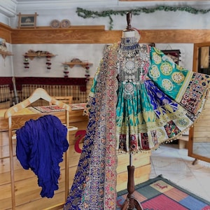 Puede incluir: Un vestido azul y verde con bordados intrincados y joyas de plata. El vestido se exhibe en un maniquí en un entorno de tienda.