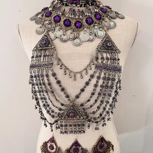 Puede incluir: Un conjunto de joyas completo con gargantilla, collar y pendientes. El conjunto incluye metal plateado con detalles morados y monedas. El collar tiene múltiples hilos y un colgante triangular. Los pendientes tienen forma de abanico.