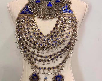 May include: An elaborate necklace with multiple layers of gold-toned chains, adorned with blue glass stones and silver-toned accents. The necklace features a choker-style collar and a central pendant with dangling elements. The design is displayed on a mannequin.