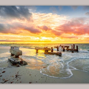 May include: A scenic sunset over a beach with a weathered wooden pier extending into the water. The sky is a vibrant mix of pink, orange, and yellow clouds. The water is a turquoise blue and the sand is white.