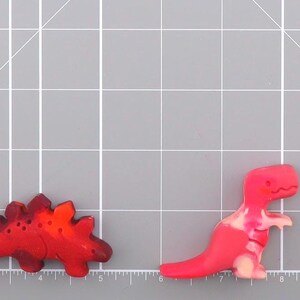 May include: Four colorful dinosaur-shaped embellishments. The dinosaurs are in shades of orange, red, and pink, with yellow and white accents. They are arranged on a gray cutting mat with grid lines. The embellishments are likely for crafts or scrapbooking.