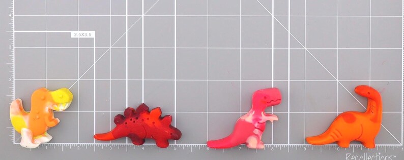 May include: Four colorful dinosaur-shaped embellishments. The dinosaurs are in shades of orange, red, and pink, with yellow and white accents. They are arranged on a gray cutting mat with grid lines. The embellishments are likely for crafts or scrapbooking.