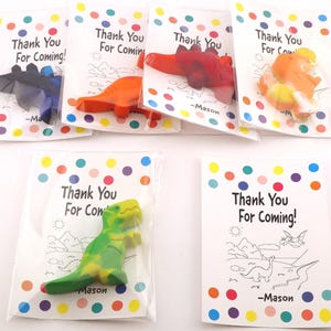 May include: A collection of dinosaur-shaped crayons in clear plastic bags, each paired with a card that reads "Thank You For Coming!" in black text. The crayons are various colors, including blue, orange, and green.