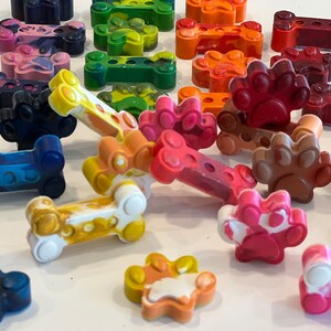 May include: A collection of colorful crayons shaped like dog bones and paw prints. The crayons are made of wax and have a variety of colors, including red, blue, green, yellow, orange, and brown.