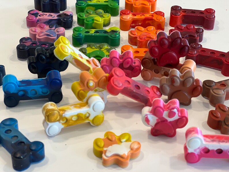 May include: A collection of colorful crayons shaped like dog bones and paw prints. The crayons are made of wax and have a variety of colors, including red, blue, green, yellow, orange, and brown.